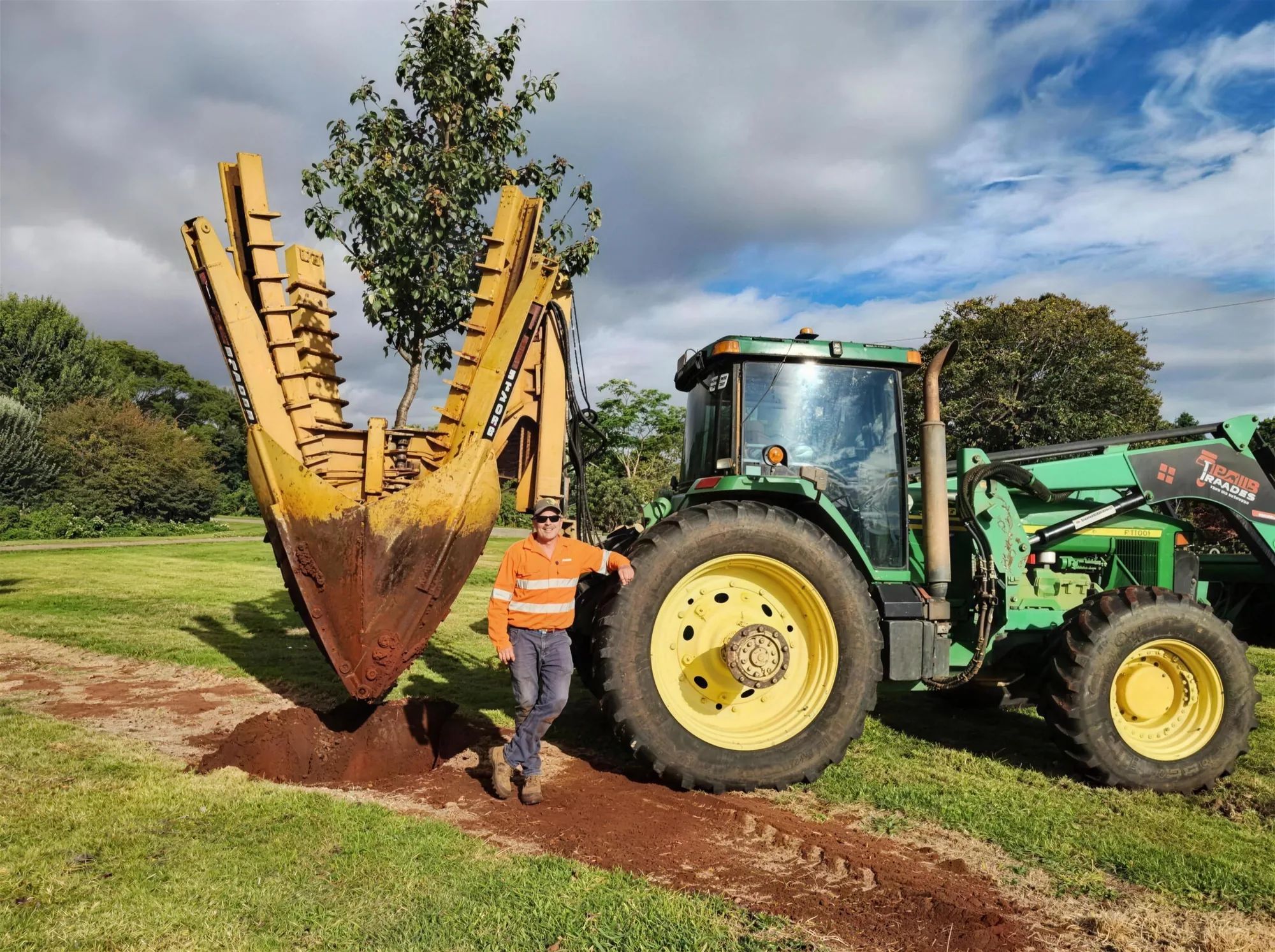 Tree spade holding a whole tree above a fresh dirt hole, Russell leaning on the tractor wheel beside it